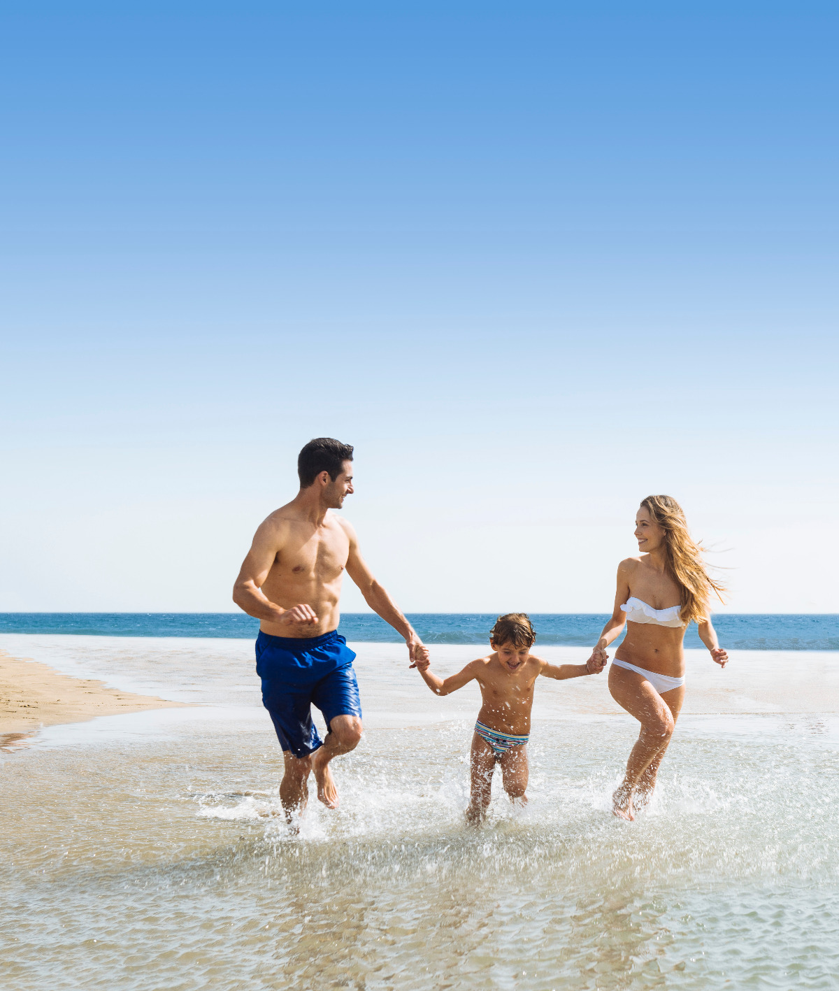 Familie mit Kind läuft lachend durch flaches Wasser am Strand, blauer Himmel und Meer im Hintergrund.